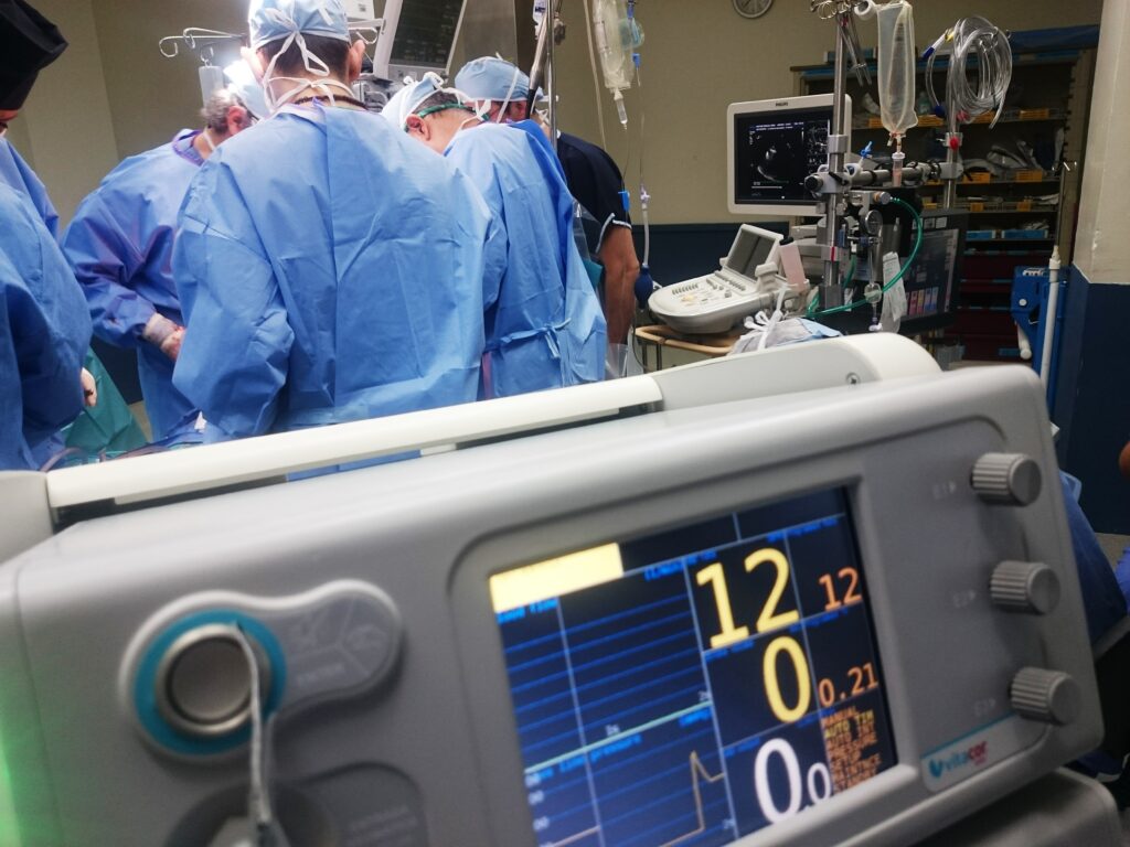 Operating theatre. Health workers in blue scrubs have their backs to the camera. In the foreground a heart monitor shows the cardiogram. Image by Natanael Melchor for Unsplash.