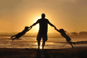 a father swinging two children by his arms on a beach during sunset
